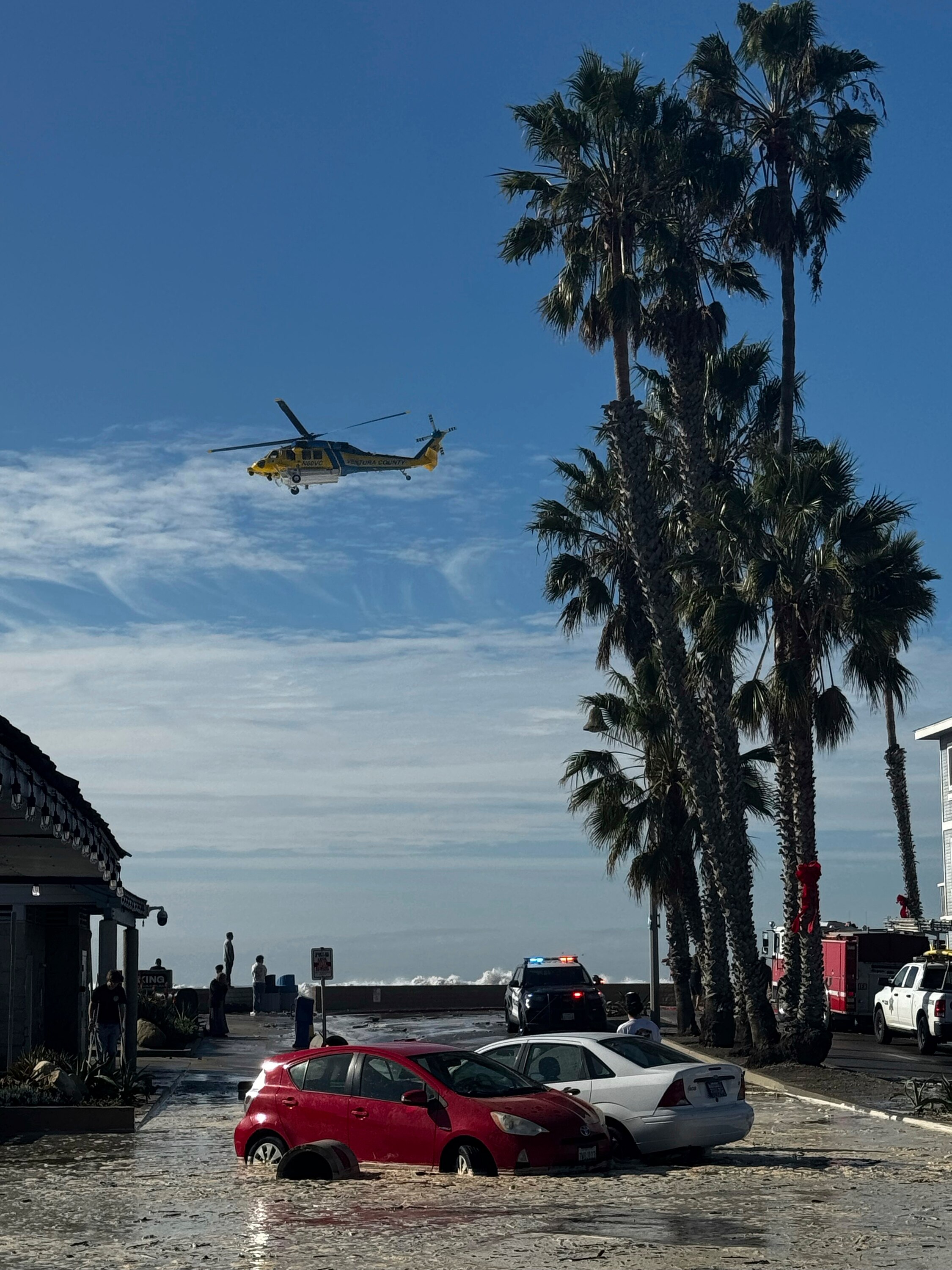 Вода высоких волн затопляет улицы.  Посреди дороги в центре фотографии стоят две затопленные машины, над ними пролетает вертолет.