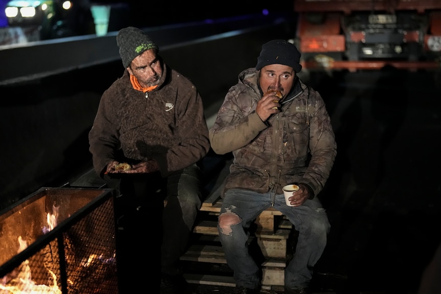 Two men sit on a road next to a fire in the day eating.