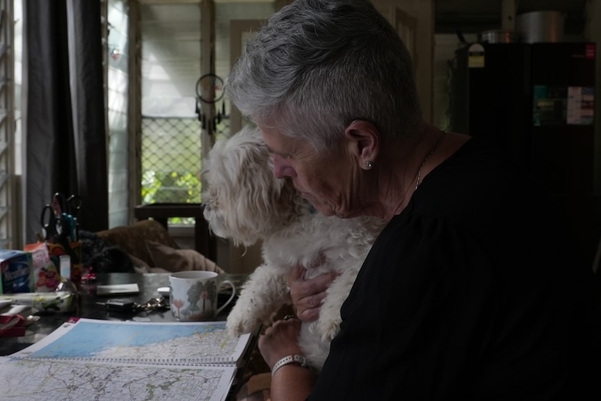 A woman with short grey hair in a black top looks at a map of Australia with her little white dog on her lap