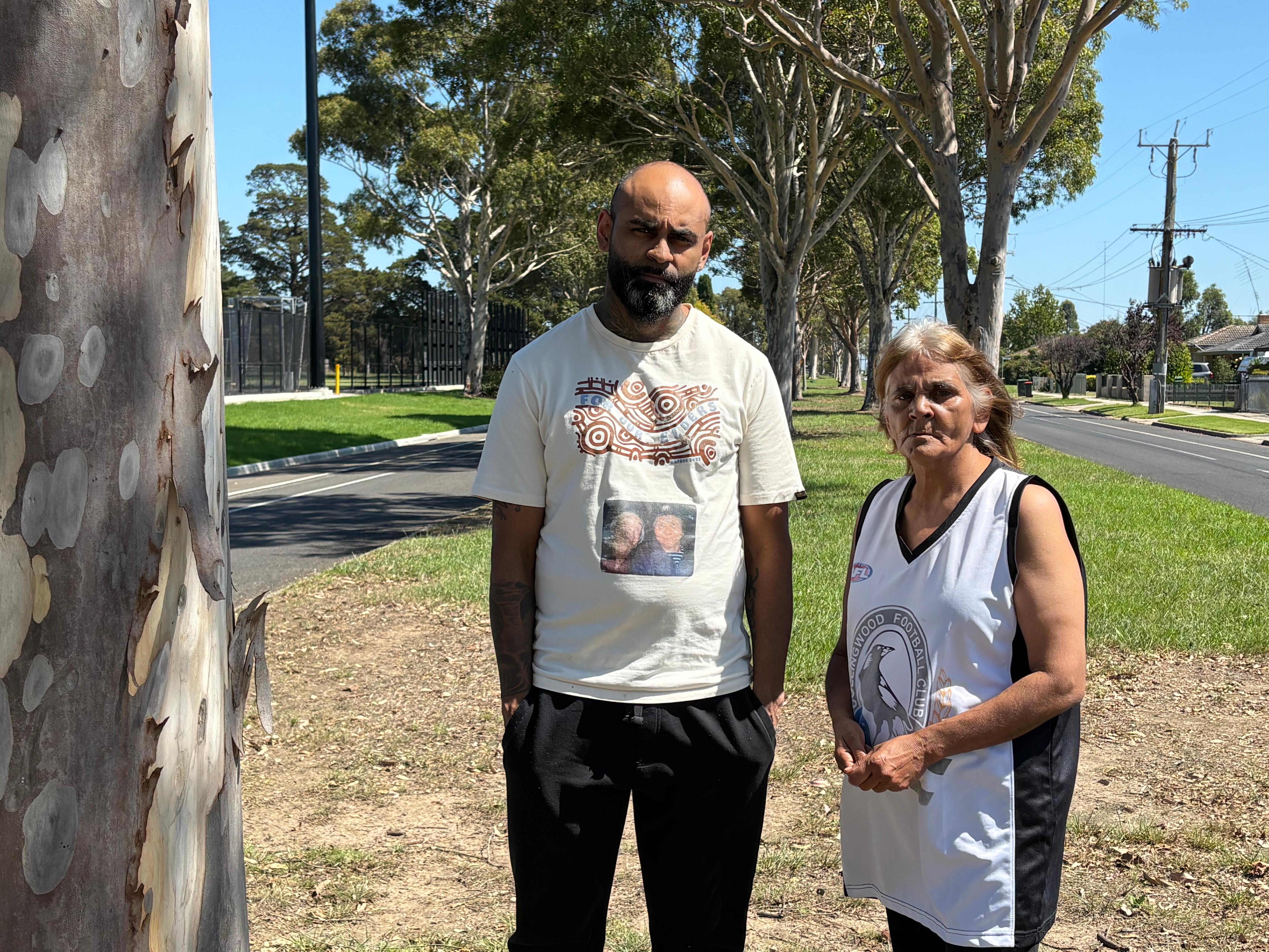Um homem indígena e sua mãe estão em um canteiro central de uma cidade regional, parecendo pensativos.
