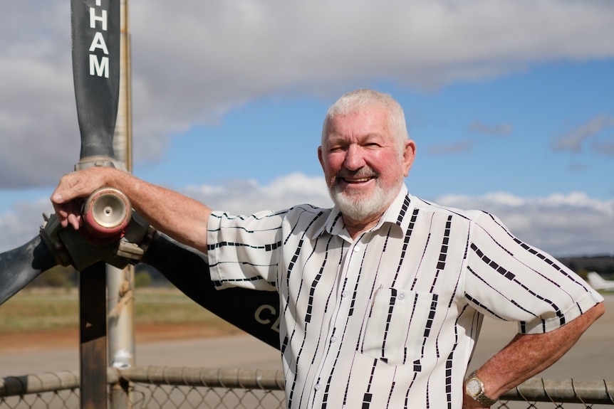 A man with grey hair and a beard standing at an aerodrome next to an old propeller blade.