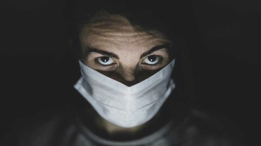 A man in a white hospital mask and dark brown eyes looks into the camera under a spotlight