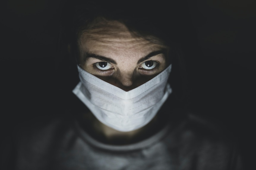 A man in a white hospital mask and dark brown eyes looks into the camera under a spotlight
