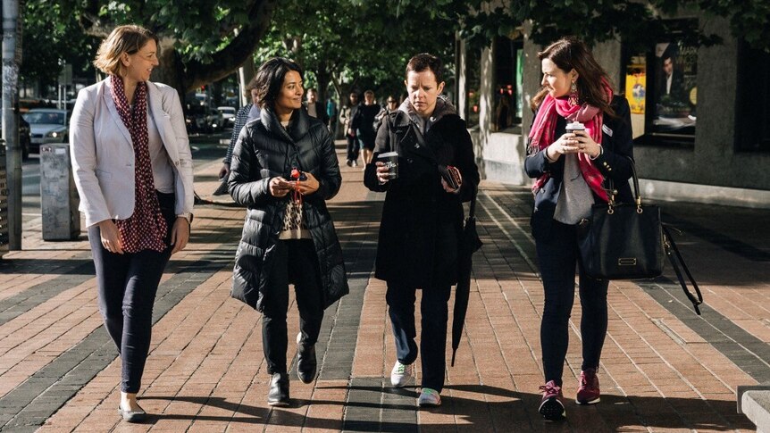 Four women walking along a street listening to one woman in the middle.