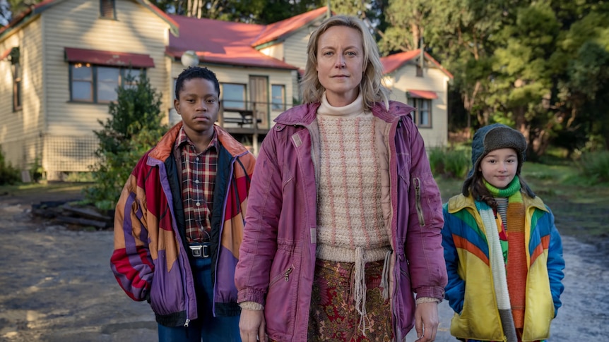 Woman in pink mauve coat with two brightly dressed children standing in front of a house