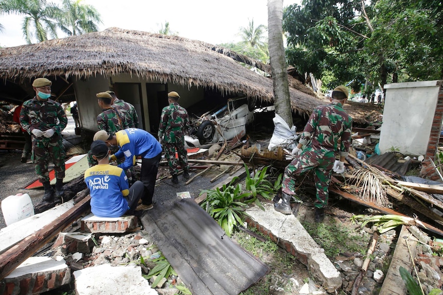 Indonesian soldiers and rescuers search for bodies buried under debris. A straw-roof house is partially collapsed on a car.