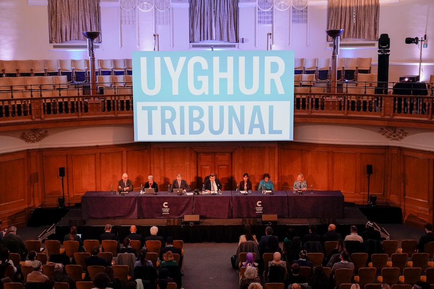 Seated panel of tribunal members with large sign behind with 