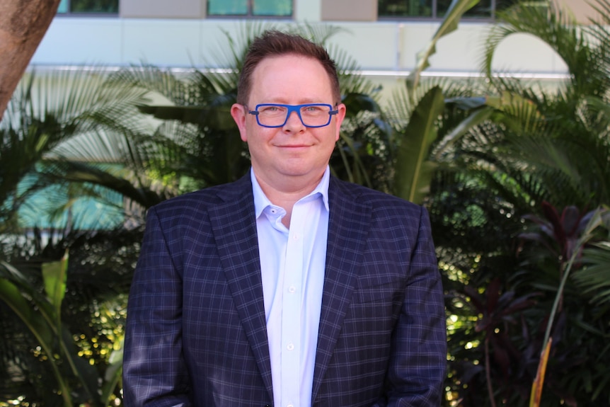 A man with a shirt, blazer and blue glasses standing in front of a fern.