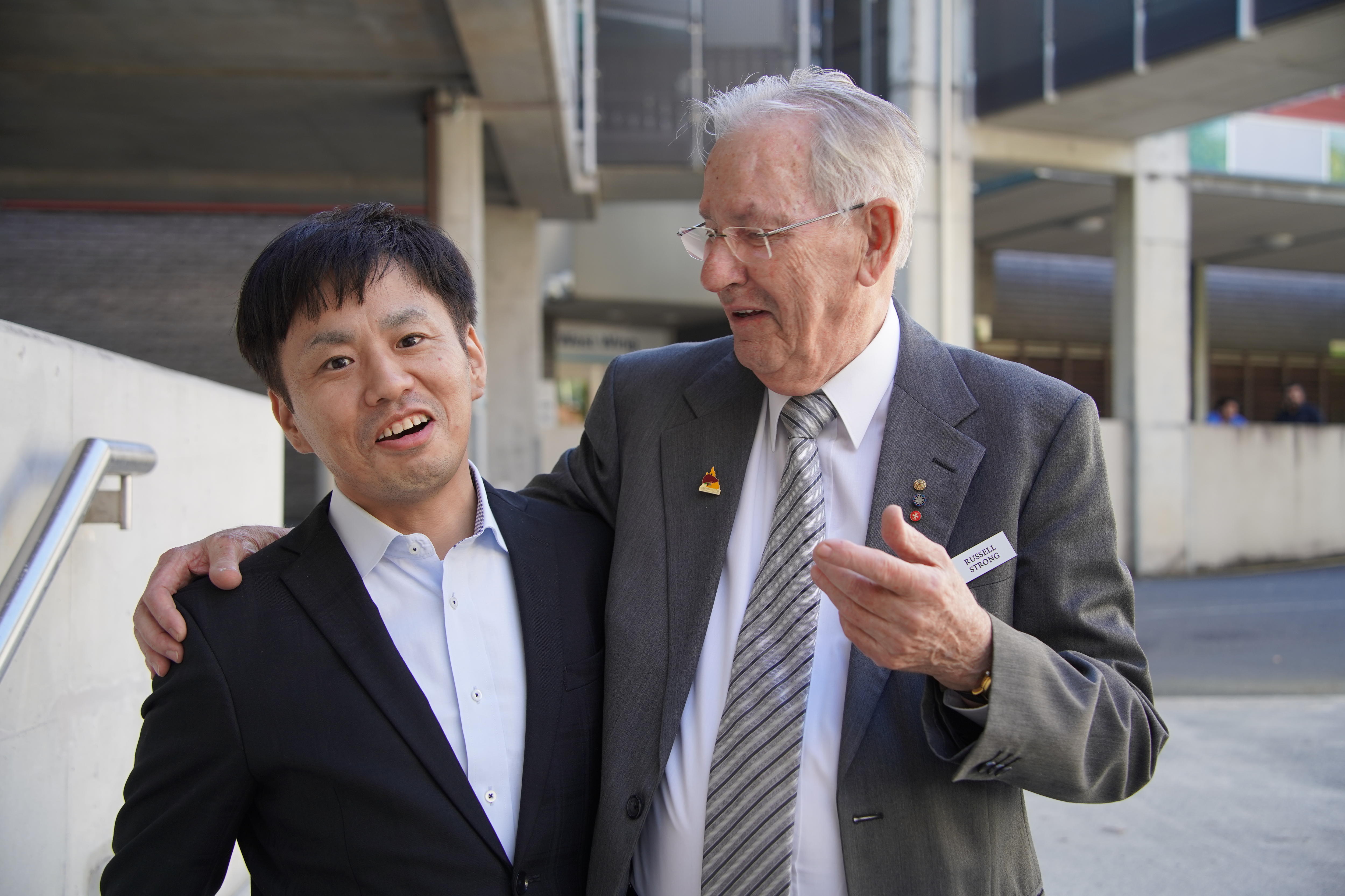 Two men in suits pose arm in arm smiling