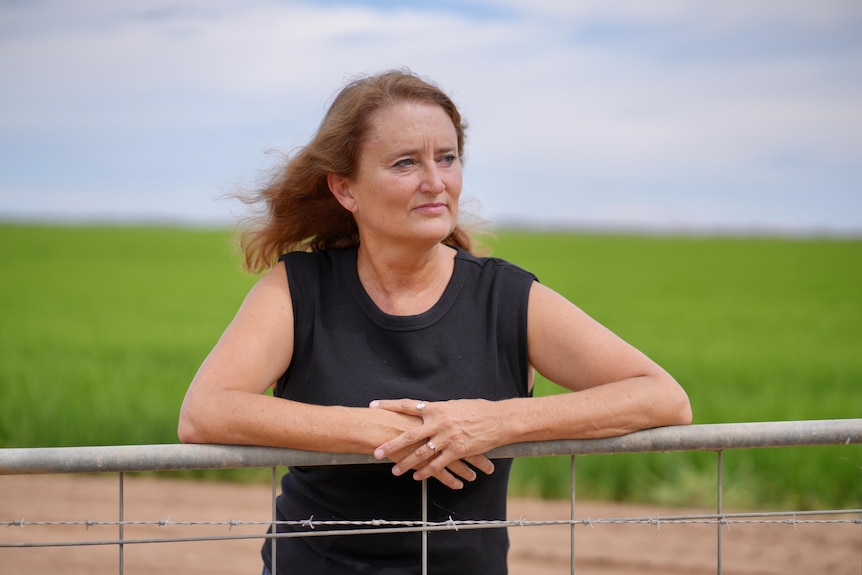 Woman with long brown hair and black tank top standing with her arms on a fence. 