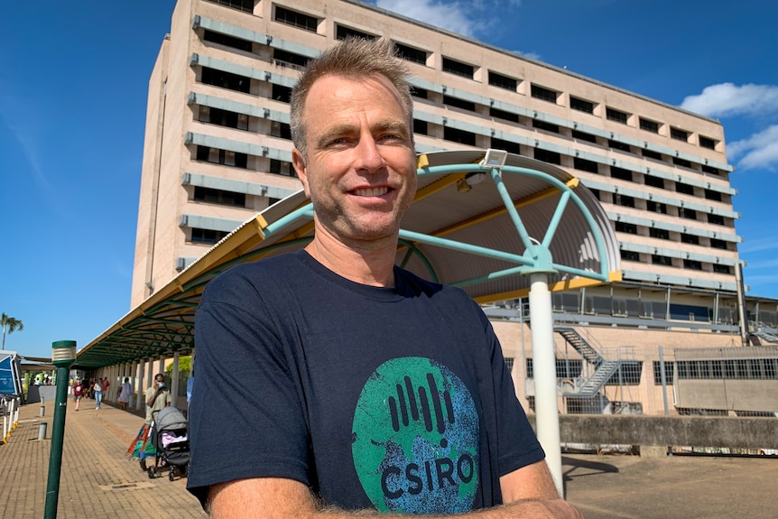 Man in CSIRO shirt arms folded sunny day large hospital in background.