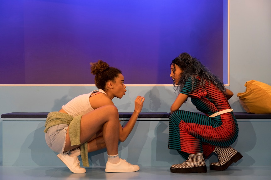 Two girls are crouched facing each other on a stage. They're having a heated conversation.