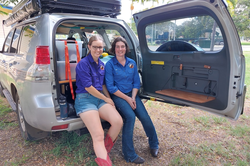 Two women in blue shirts sit on the tailgate of a four wheel drive, the door is open and the car is full of boxes.