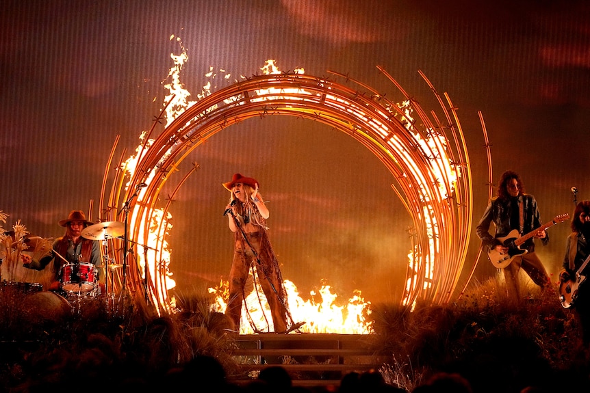 A young woman in a cowboy hat sings into a microphone osntage in front of a flaming circle.