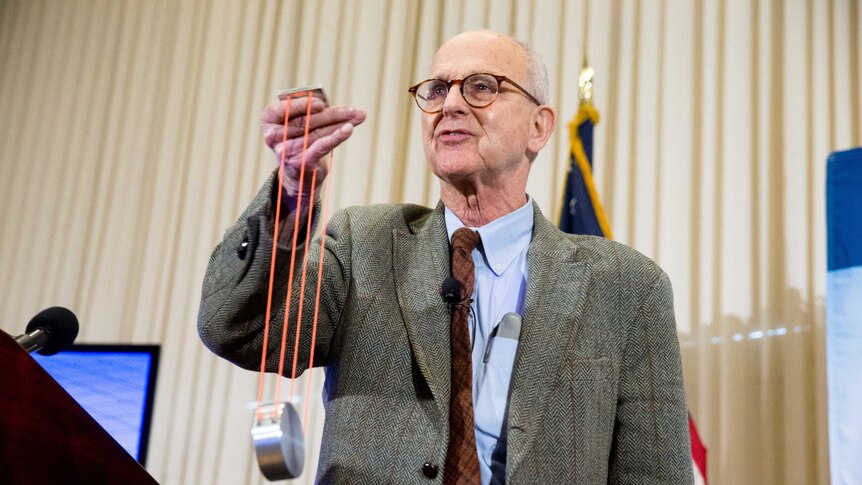 Rainer Weiss holds a piece of equipment that looks like a metal cylinder attached to orange strings.