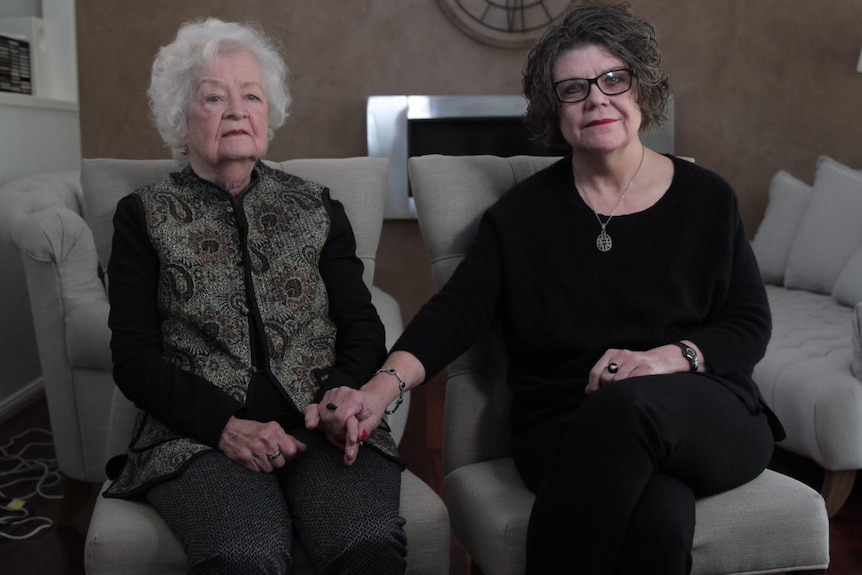 Beryl Cunnane and her daughter Mardella Bassett sitting on sofa chairs holding hands