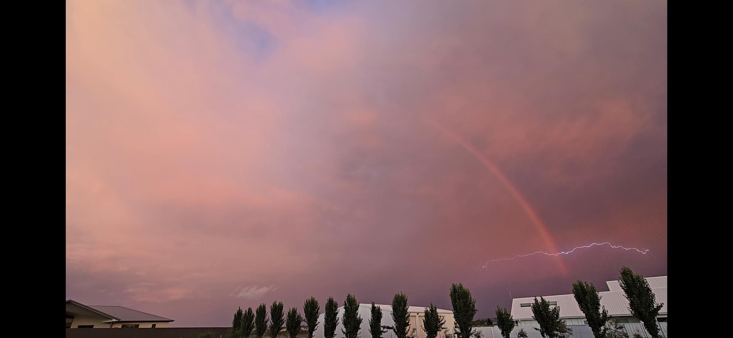 A bolt of lightning shoots across the sky in front of a partial rainbow arc