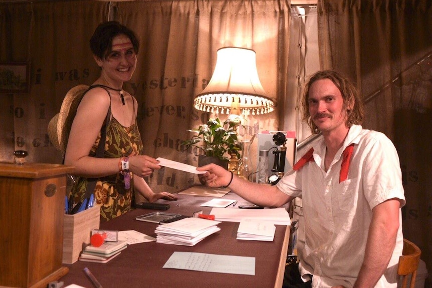 Woman hands letter to man in room with hessian walls