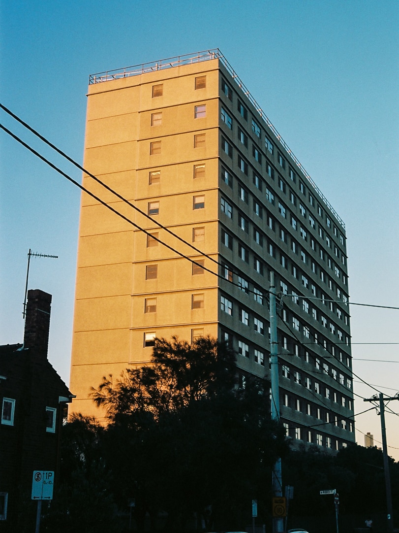 On a bright blue day at sunset, you view a public housing tower bathed in golden light,