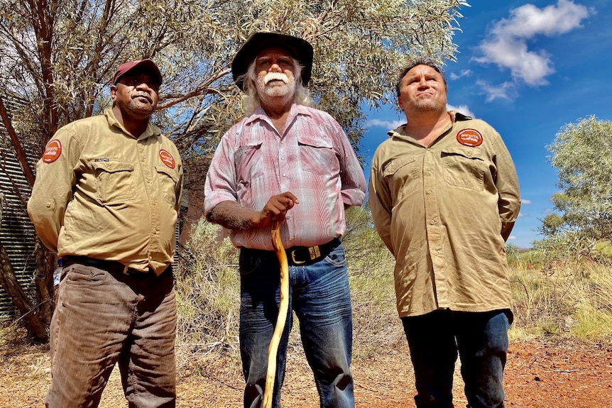 A senior Aboriginal man with a bushy white moustache and broad hat stands beneath a tree with two men in ranger shirts.