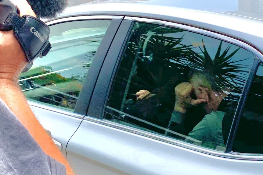 A man with his head down in the back of a police car on a driveway