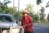 A man in a red button-up shirt and straw hat leans on his ute.