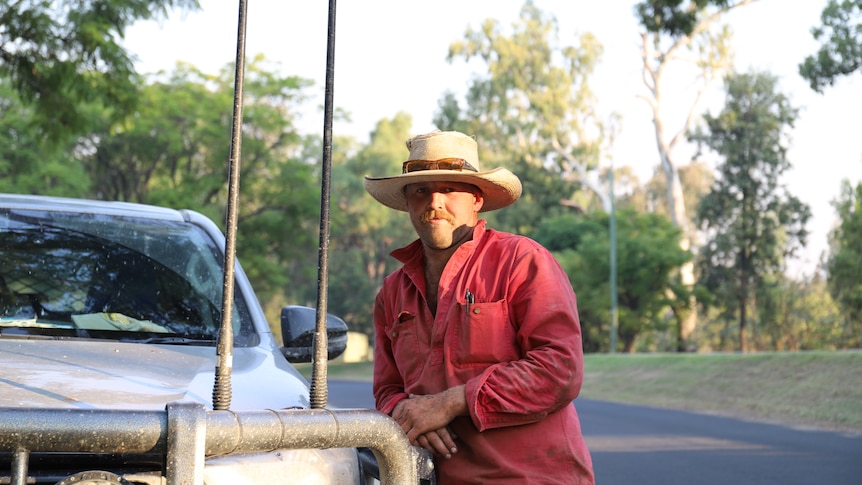 A man in a red button-up shirt and straw hat leans on his ute.