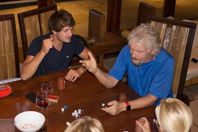 Richard Branson sits at a table covered in wine and dice with members of his staff.