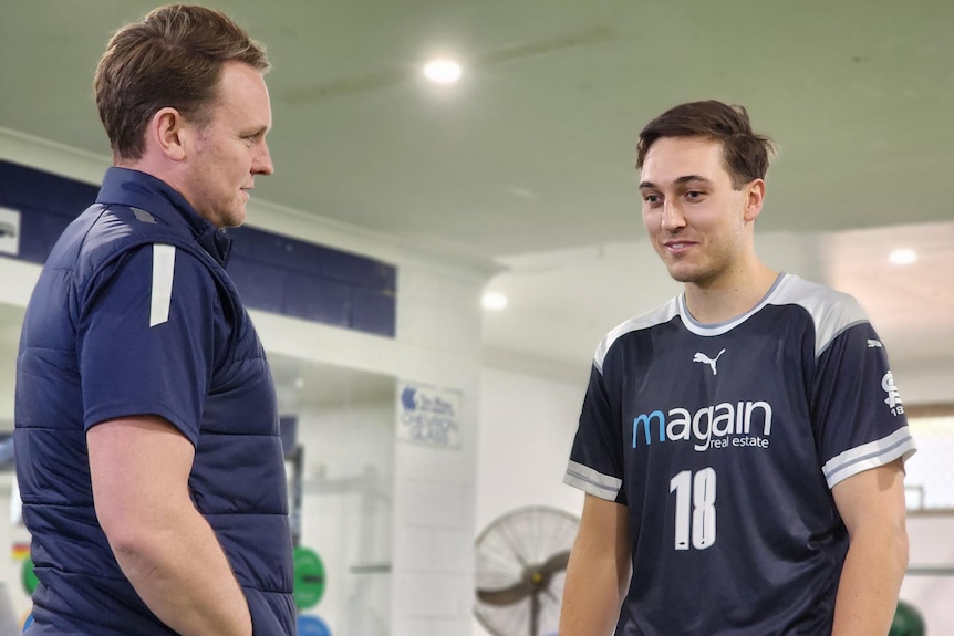 Two men in blue tops talking to each other in a gym.