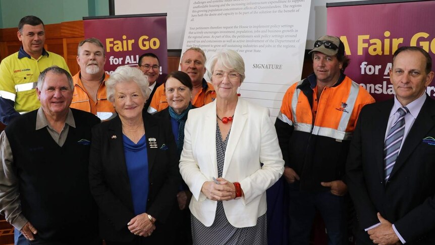 A group of local government representatives stand in front of a poster and banners stating "Fair Go for All Queenslanders".