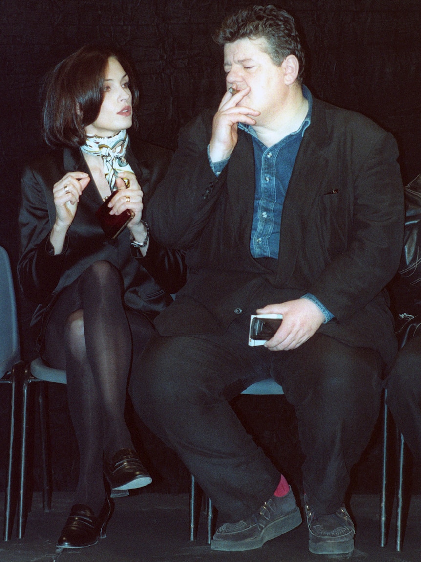 coltrane looks into the distance while smoking a cigar as janssen speaks to him, both on plastic folded chairs