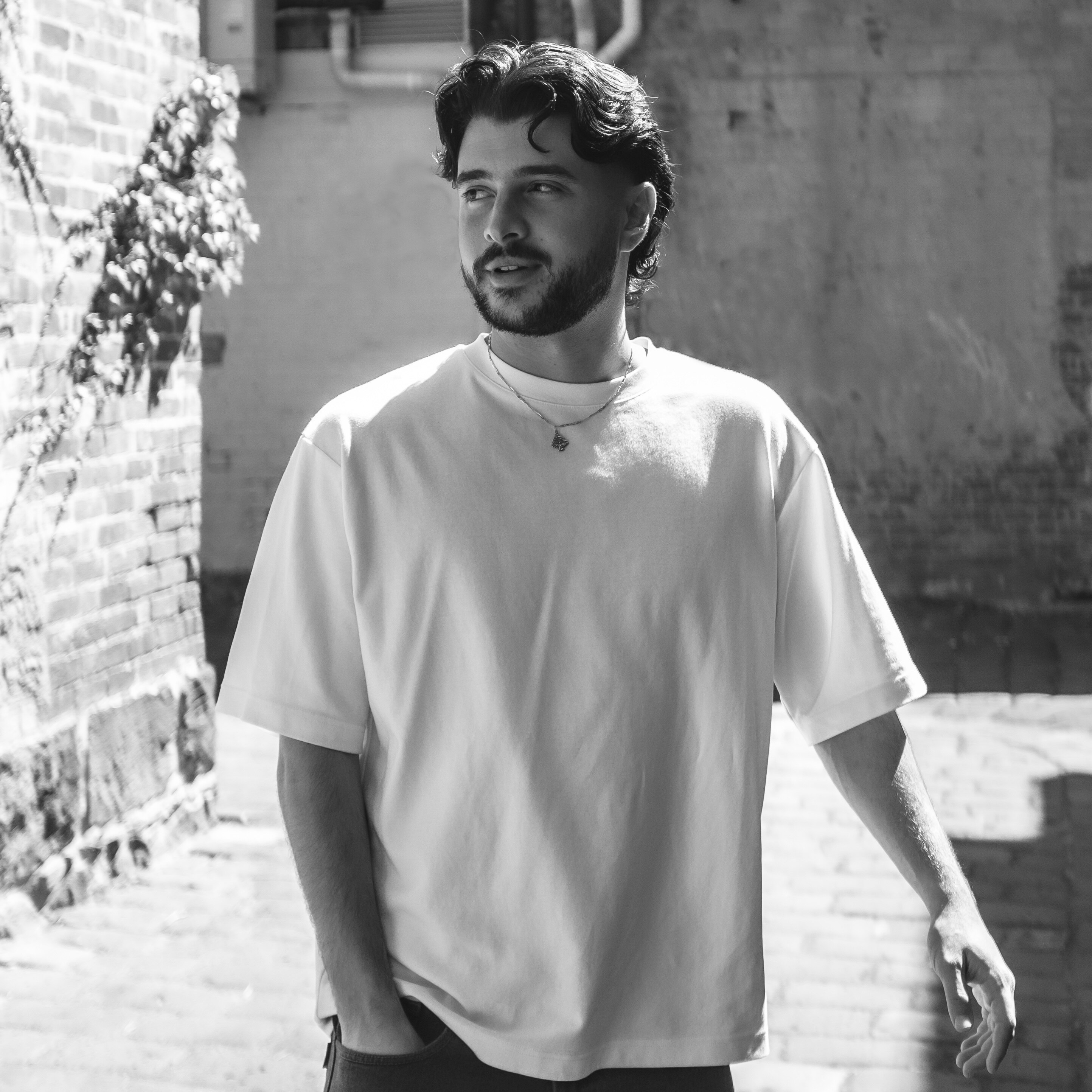A black and white photo of a guy in his 20s with a white t-shirt. He's looking to the side on a stoney street.