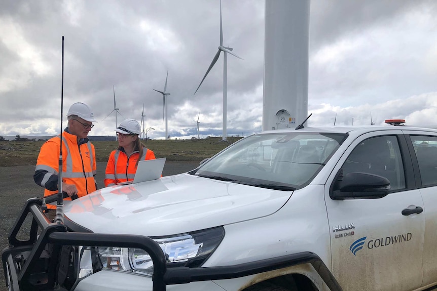 A man and a woman in orange high-vis stand next to a car in front of some large wind turbines.