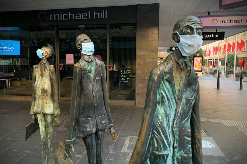 Three bronze sculptures of men in business suits near a Melbourne mall, each wearing a blue surgical mask.