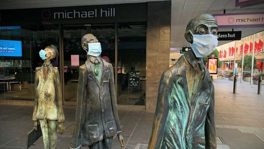 Three bronze sculptures of men in business suits near a Melbourne mall, each wearing a blue surgical mask.