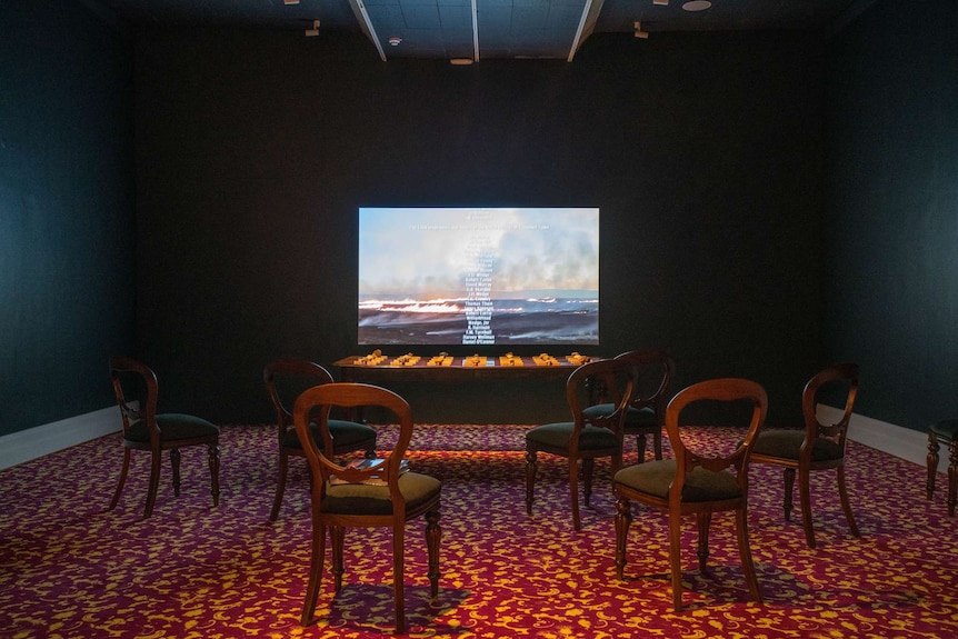 Gallery room with red and yellow patterned carpet, video screen, and dining table and chairs laid out in front.