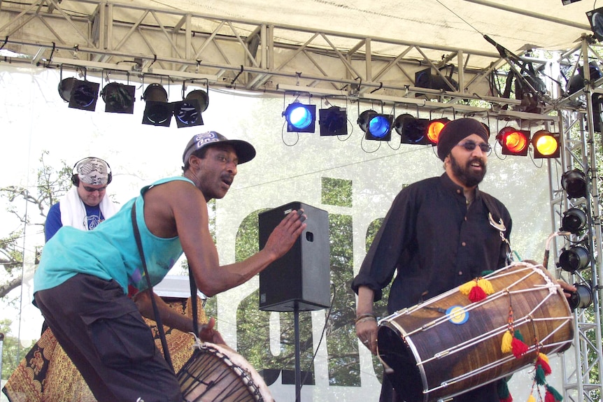 Three men play percussion on an outoor stage during the day