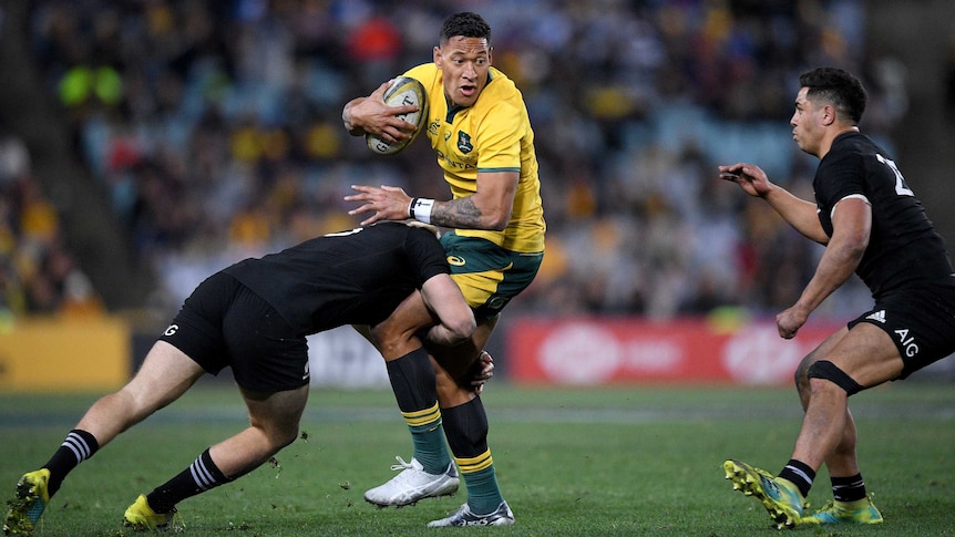 A rugby player wearing black tackles a players wearing yellow around the hips whilst another player in black watches on