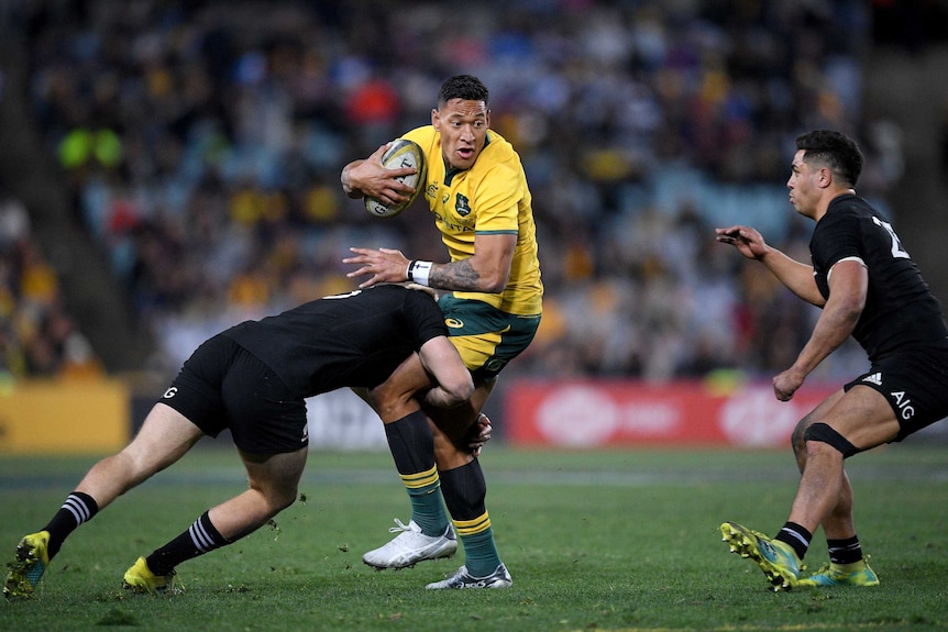 A rugby player wearing black tackles a players wearing yellow around the hips whilst another player in black watches on