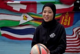 A woman in a navy blue soccer top and wearing a head scarf, holds a white, black and orange striped soccer ball