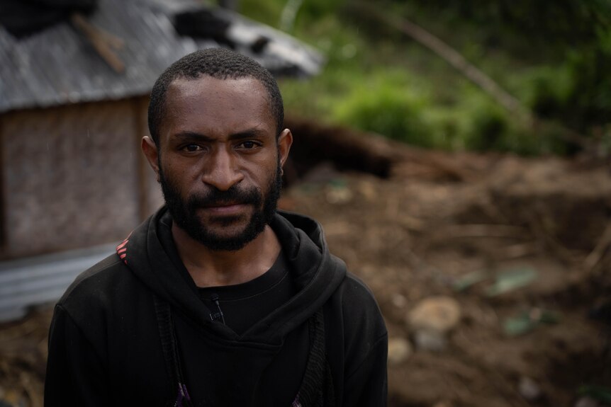 A young Papua New Guinean man in a black hoodie looks directly at the camera in front of a shack in the forest.