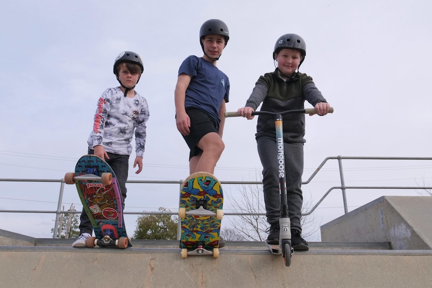 Three kids about to skate or scooter down a half-pipe structure