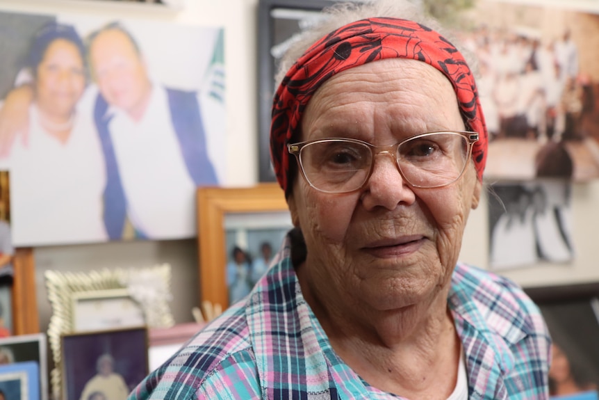 A woman in a red headscarf and glasses stands in front of family photos