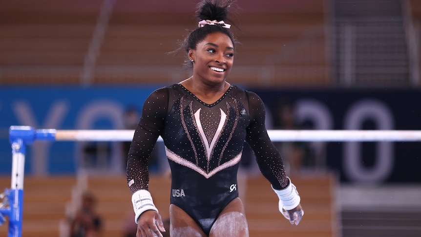 gymnast simone biles walks away from the uneven bars smiling and swinging her arms. she has chalk on her legs