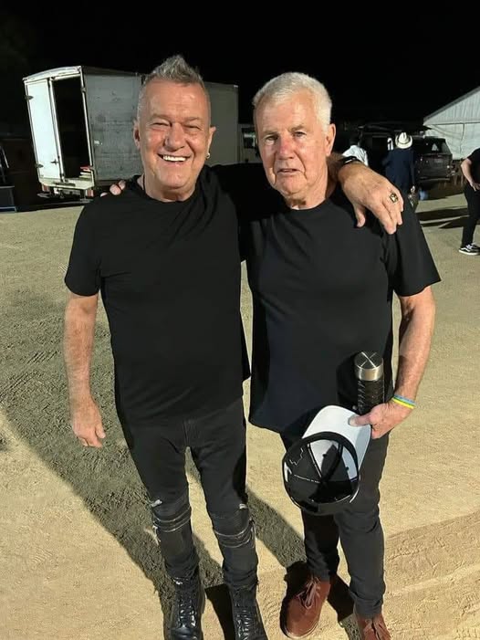 Two grey haired men in black shirts and black jeans stand arm in arm near a truck at night.