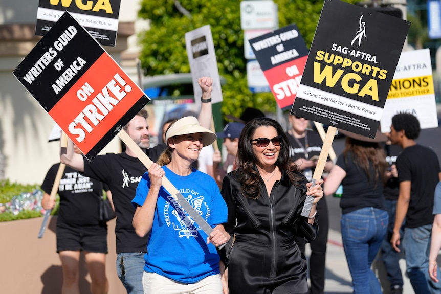 Fran Drescher in black velvet tracksuit and Meredith Stiehm in blue T smile while holding protest signs