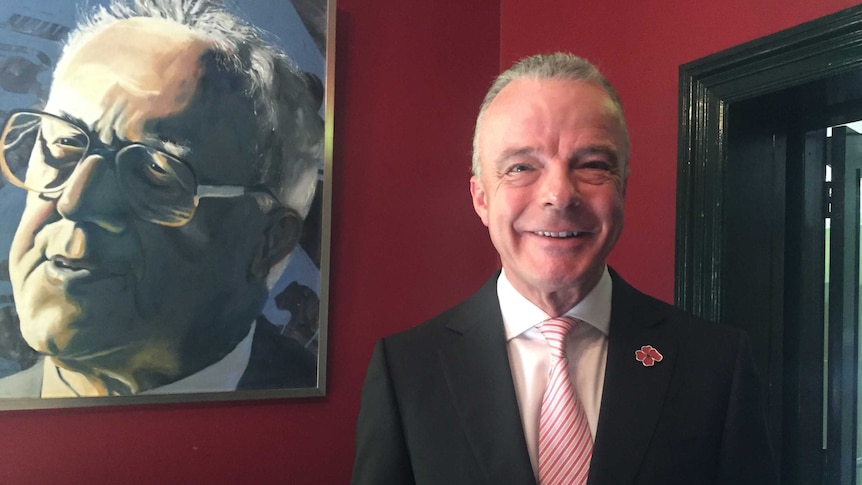 Director of the Australian War Memorial, Dr Brendan Nelson, stands in front of a portrait painting in a red room.