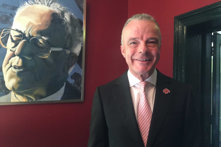 Director of the Australian War Memorial, Dr Brendan Nelson, stands in front of a portrait painting in a red room.