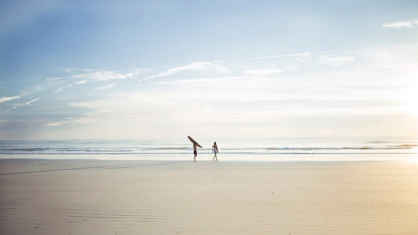 Two people carrying surfboards walk towards a calm ocean.