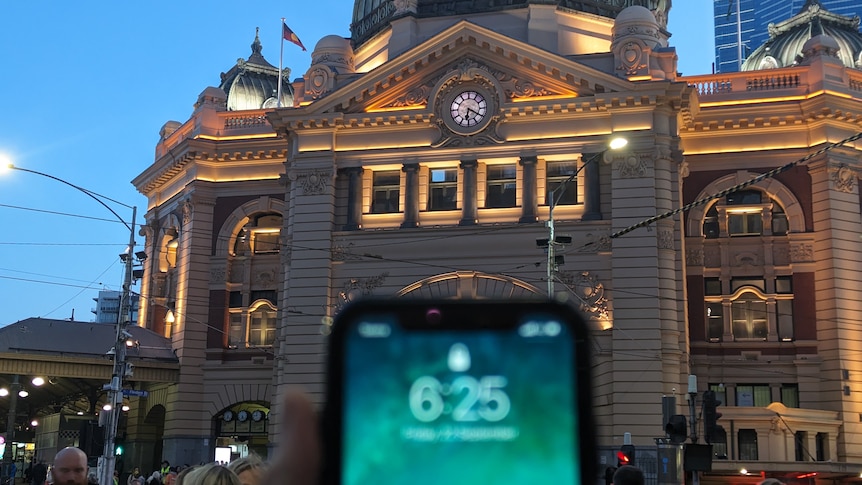 A phone screen showing the time 6.25 is in front of the analog flinders st station clock showing 6.20